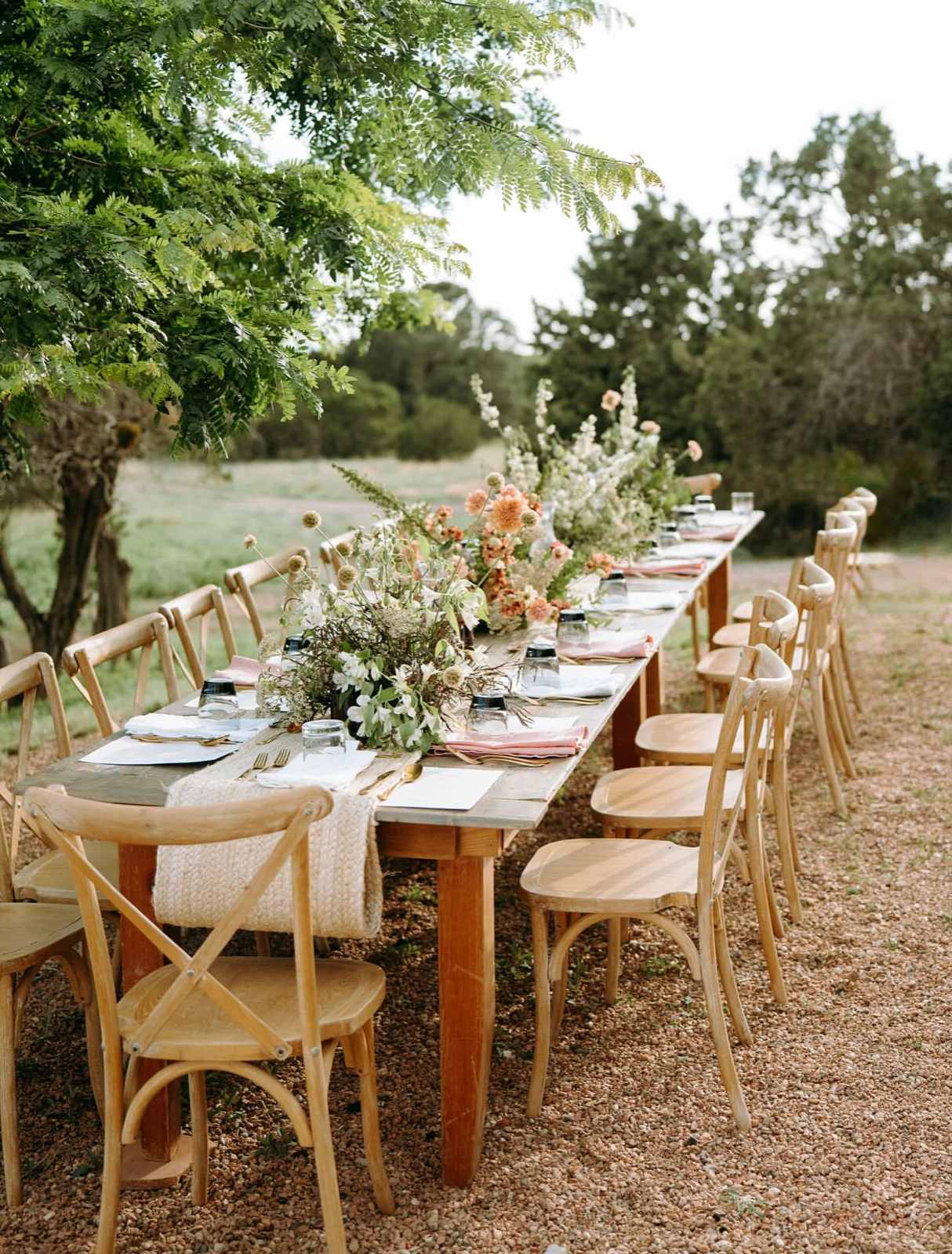 A long outdoor farm table set with florals, gold flatware and cross-back chairs under a tree.