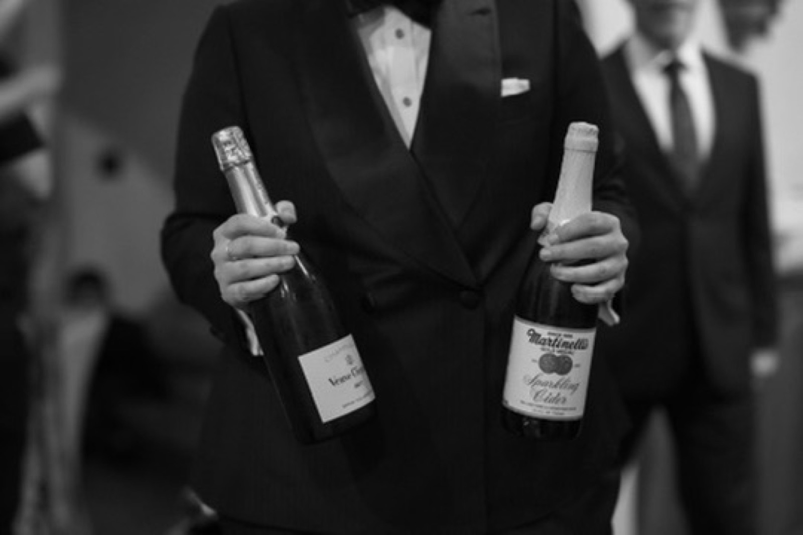 A guest in a tuxedo holding champagne and sparkling cider at a black-tie Net Zero Ranch event, in black and white.