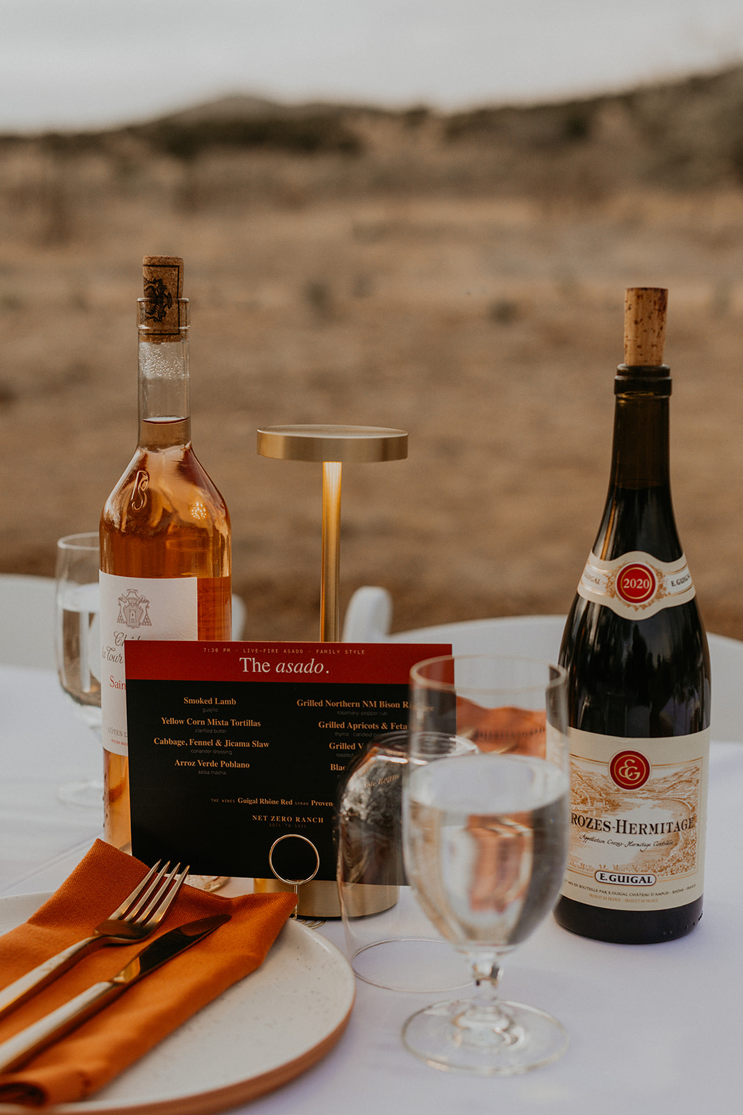 The Asado menu card and wines on the table at dusk, ranch beyond — The Asado table.