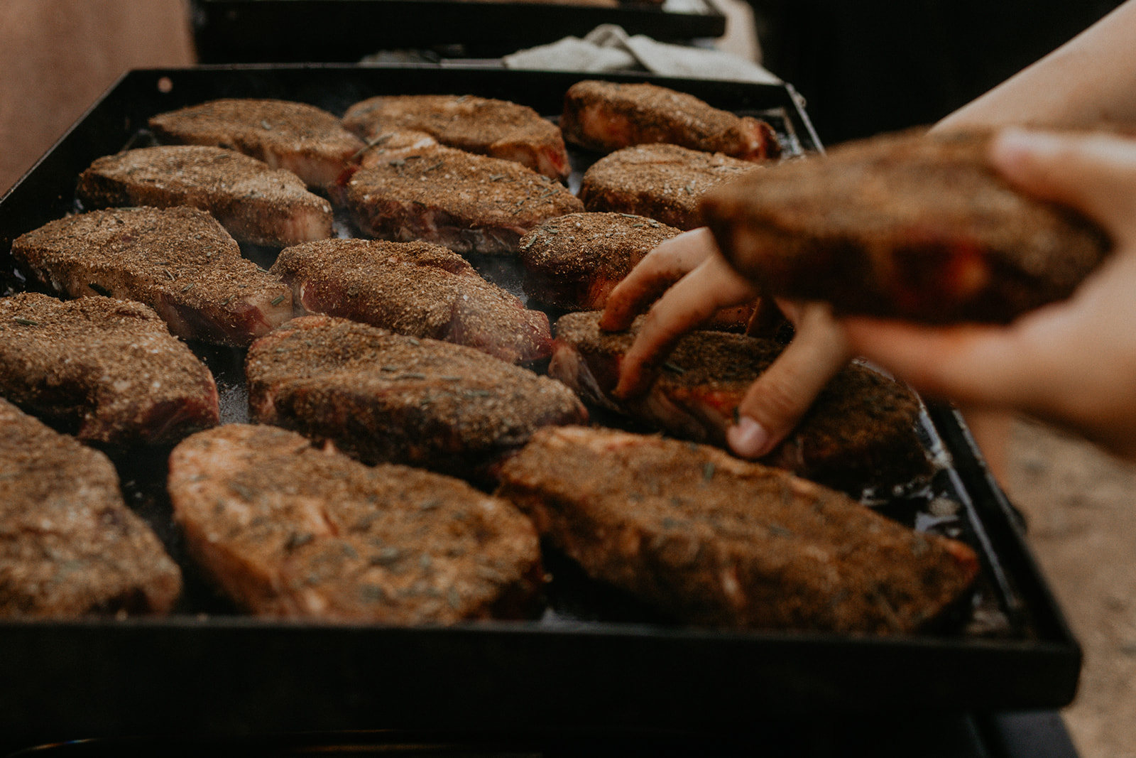 Hands laying spice-crusted bison ribeyes onto the plancha — The Asado table.