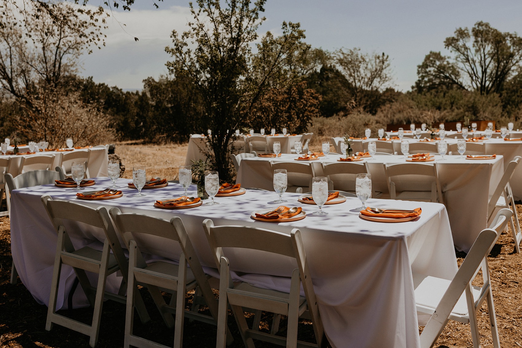 Outdoor reception tables set with white linen, rust napkins and gold flatware in the Northern New Mexico landscape.